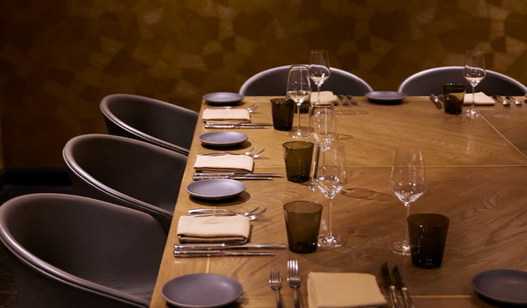 A wooden dining table set with plates, folded napkins, cutlery, and wine glasses arranged neatly, surrounded by upholstered chairs in a dimly lit restaurant interior.